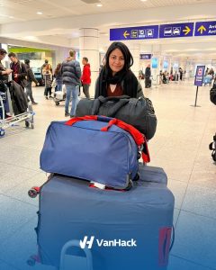 Aashi is the airport pushing her luggage. Many blue big heavy suitcases. She is has long black hair and is smiling