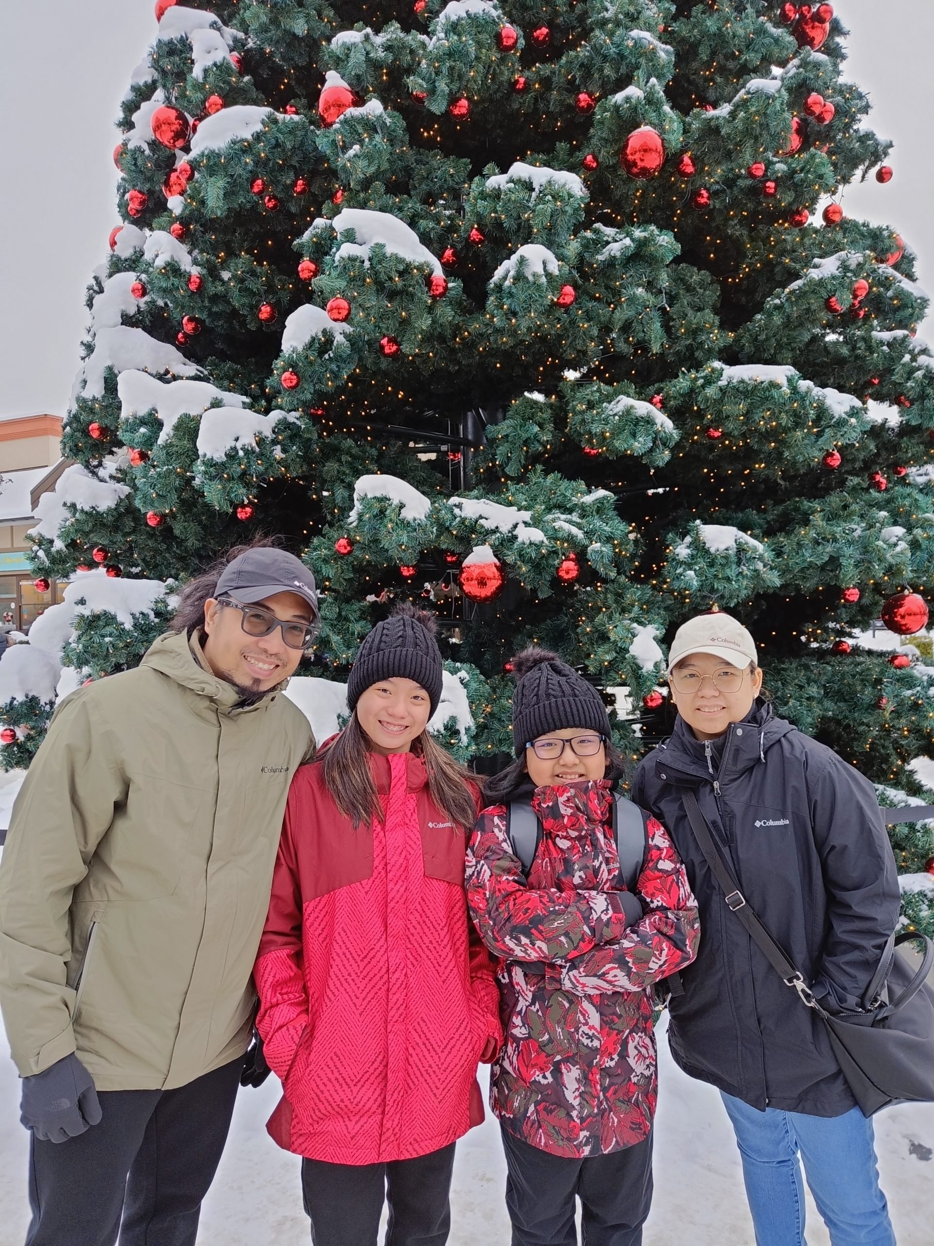 Jesper is posing with his family in front of a big Christmas tree outside. They are wearing heavy winter clothes, the ground and the tree are covered in snow.
