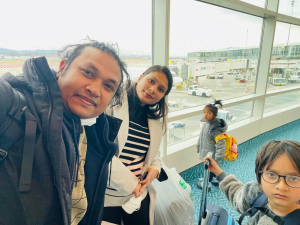 Tomas is taking a selfie with his family at the airport. He is a young man in a blue heavy coat. His wife is standing next to him. She has long dark hair and is wearing a white coat. Their kids are standing with them. The boy wear glasses and is look at the camera. The girls is behind wearing a gray coat.