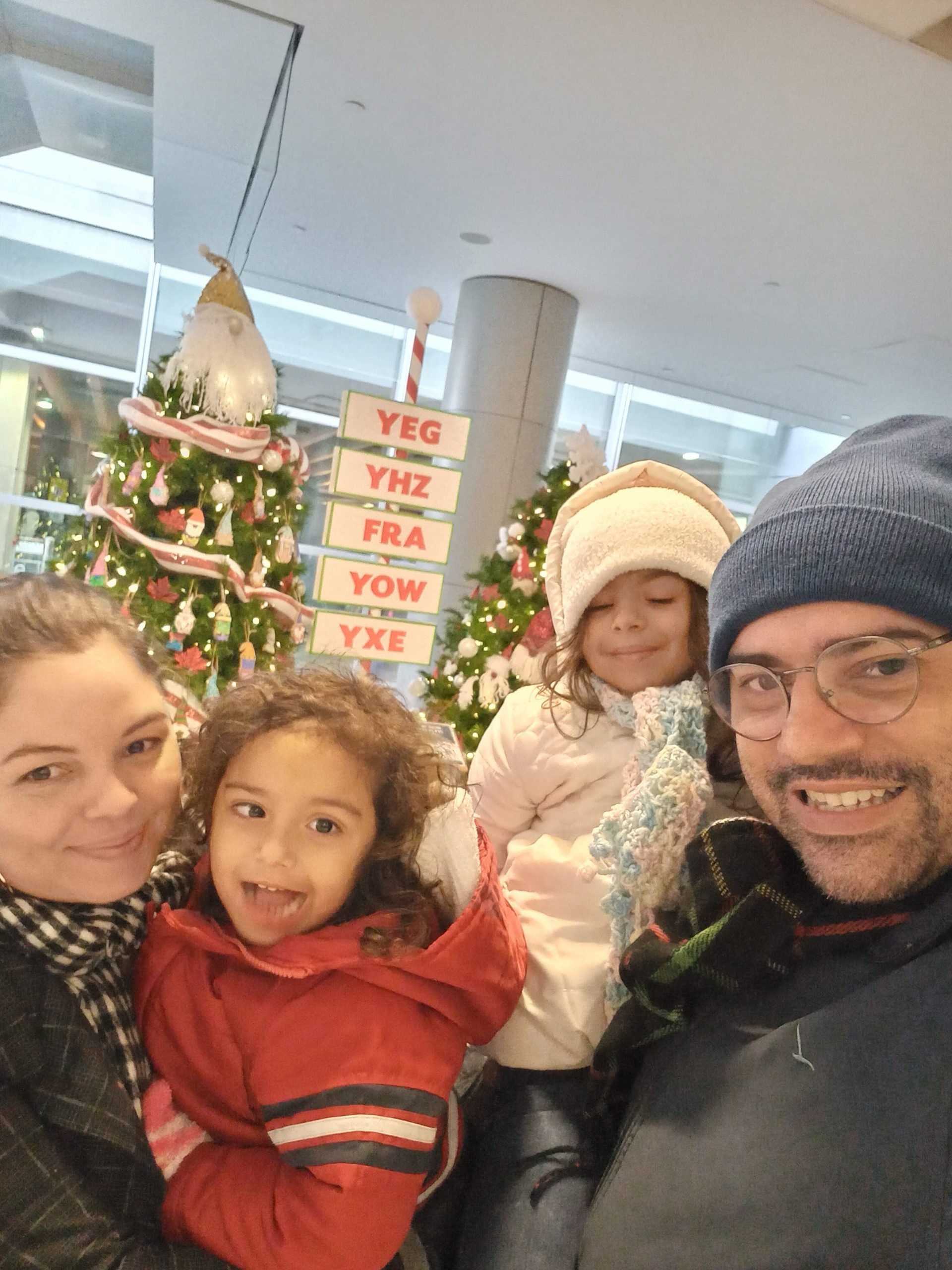 Edinor and his family pose together in front of a small Christmas tree. His family is a wife and two little girls and they are wearing heavy winter clothes